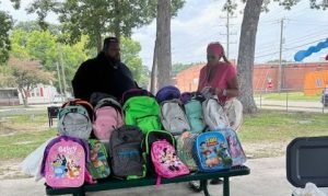 Howard and a community volunteer standing behind a table filled with backpacks collected for local students.